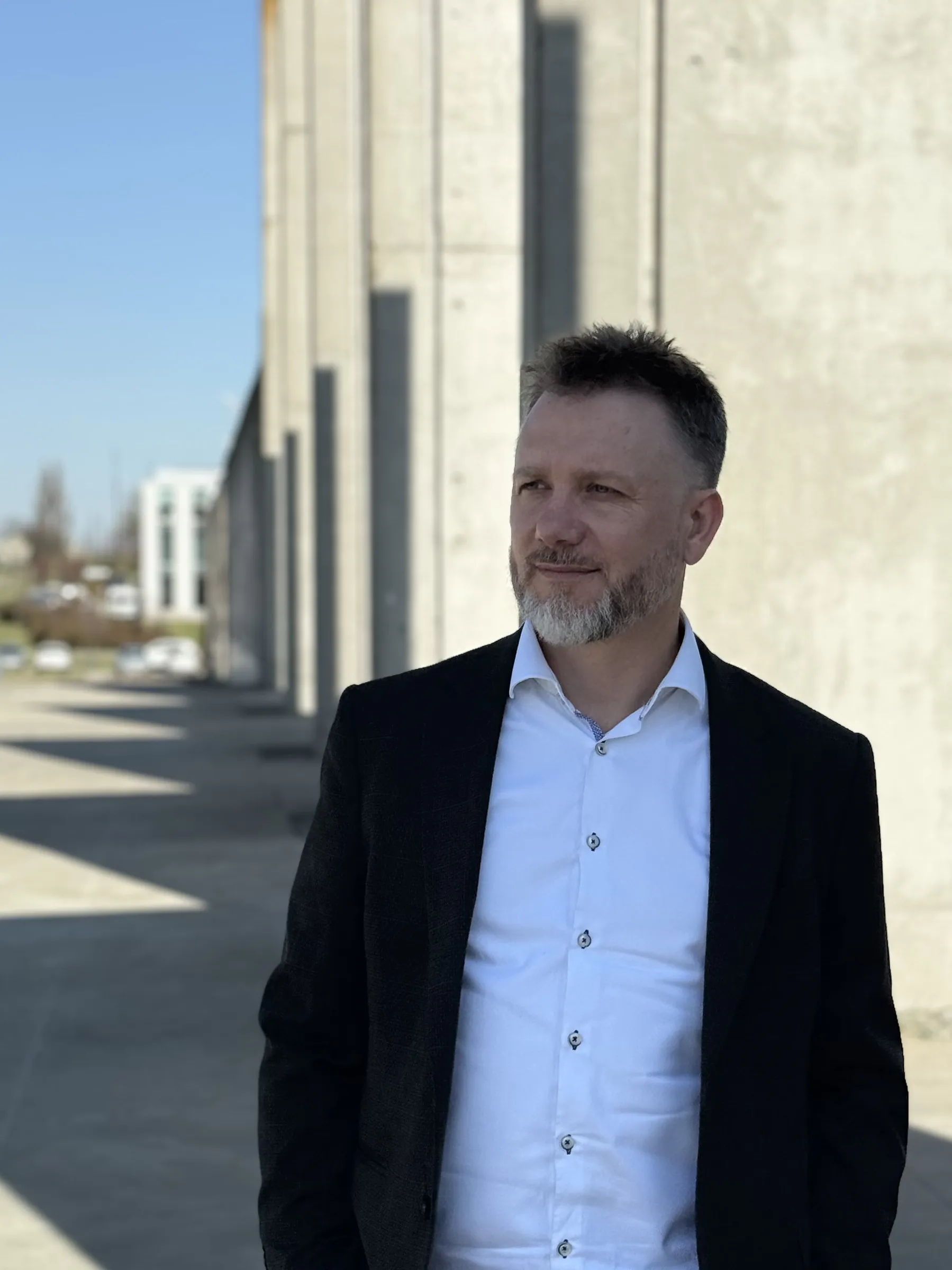 Piotr Kruk przed budynkiem o betonowej elewacji, w ciemnej marynarce i białej koszuli