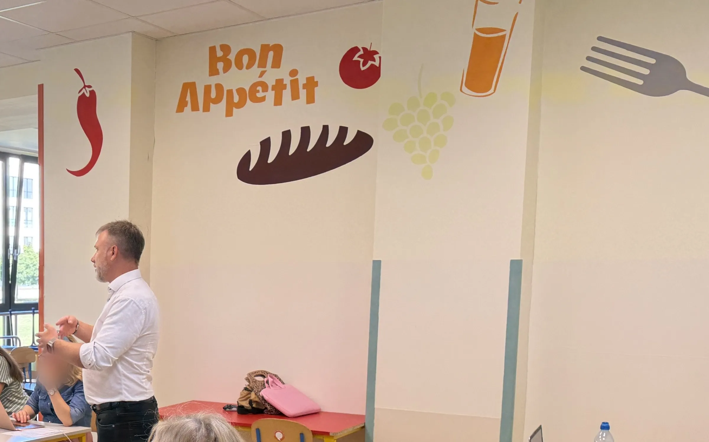 Piotr Kruk w białej koszuli rozmawia z uczestnikami szkolenia w szkolnej jadalni; na ścianie mural z napisem „Bon Appétit”, chlebem, owocami i widelcem