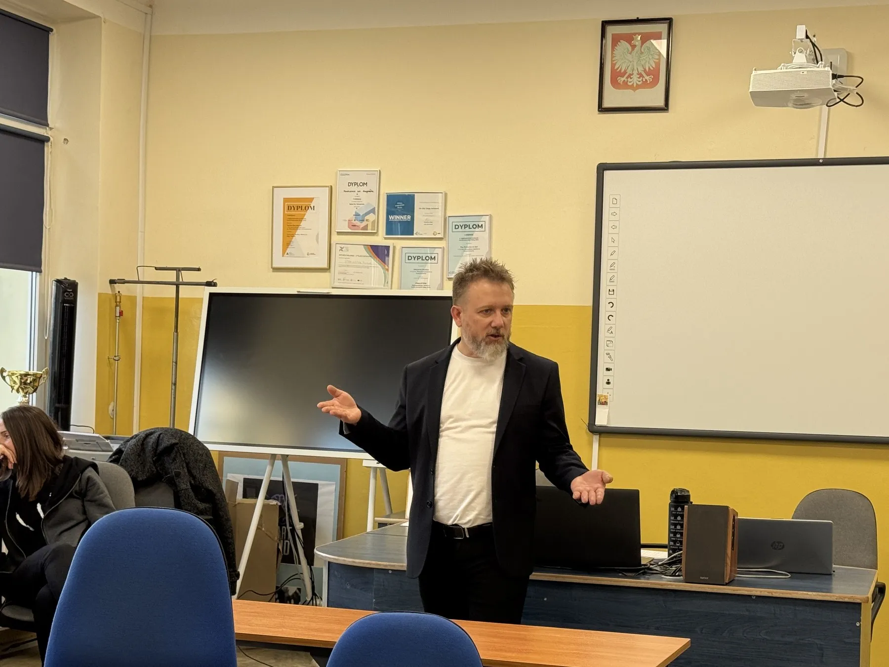 Piotr Kruk tłumaczy coś w sali lekcyjnej, gestykulując obiema rękami przy tablicy