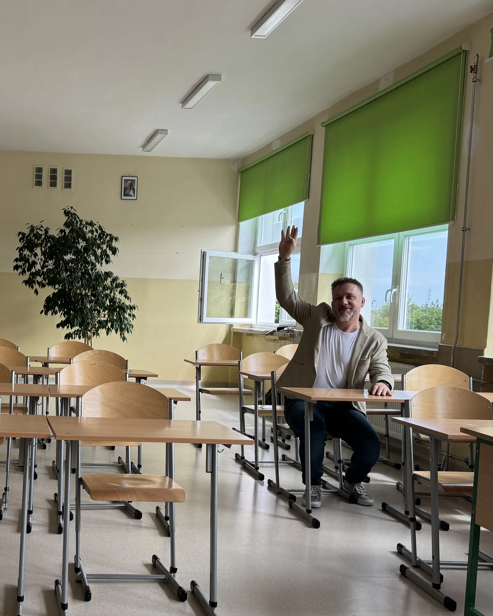 Piotr Kruk siedzi w ławce w pustej szkolnej klasie i podnosi rękę jak uczeń, który się zgłasza