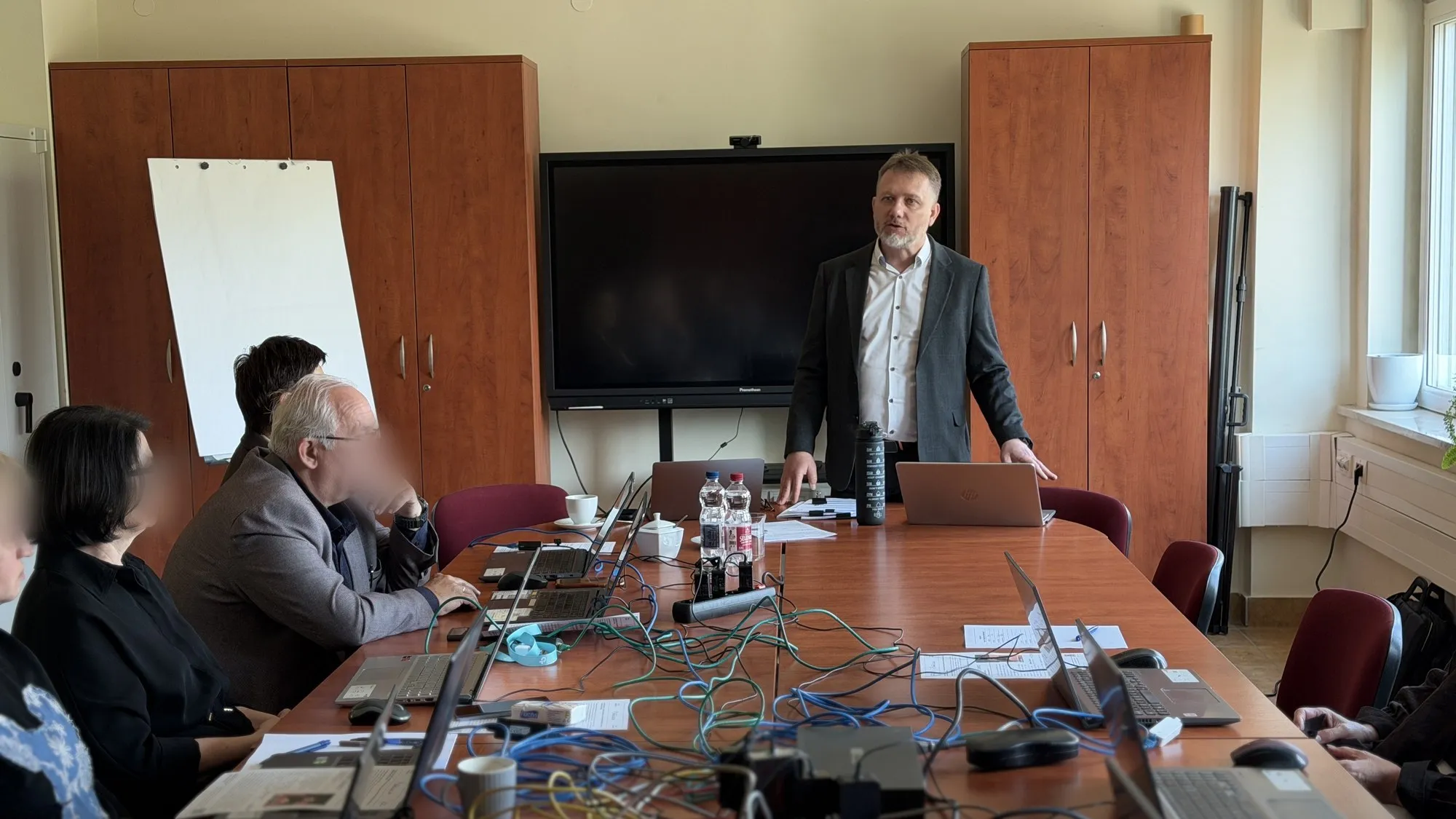 Narada przy długim stole zastawionym laptopami i kablami; prowadzący stoi przy ekranie