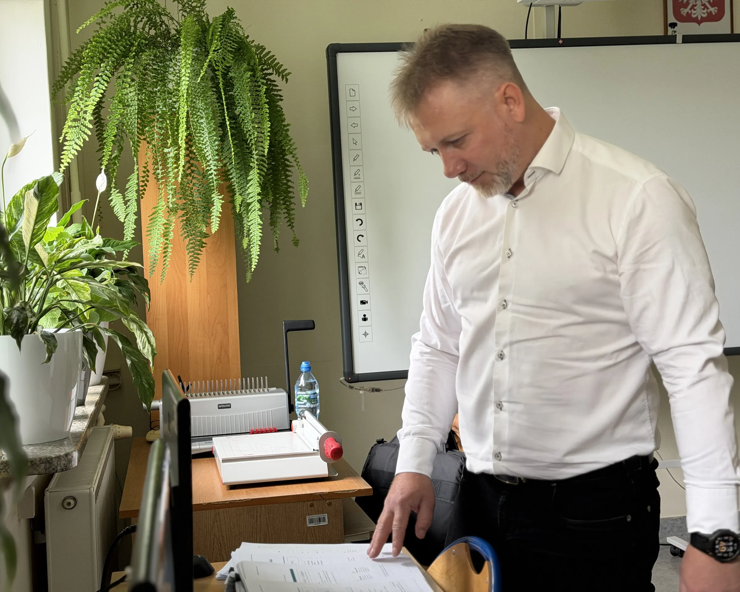 Piotr Kruk przegląda plik wydruków przy biurku w szkolnej pracowni; obok bindownica, paproć i tablica interaktywna