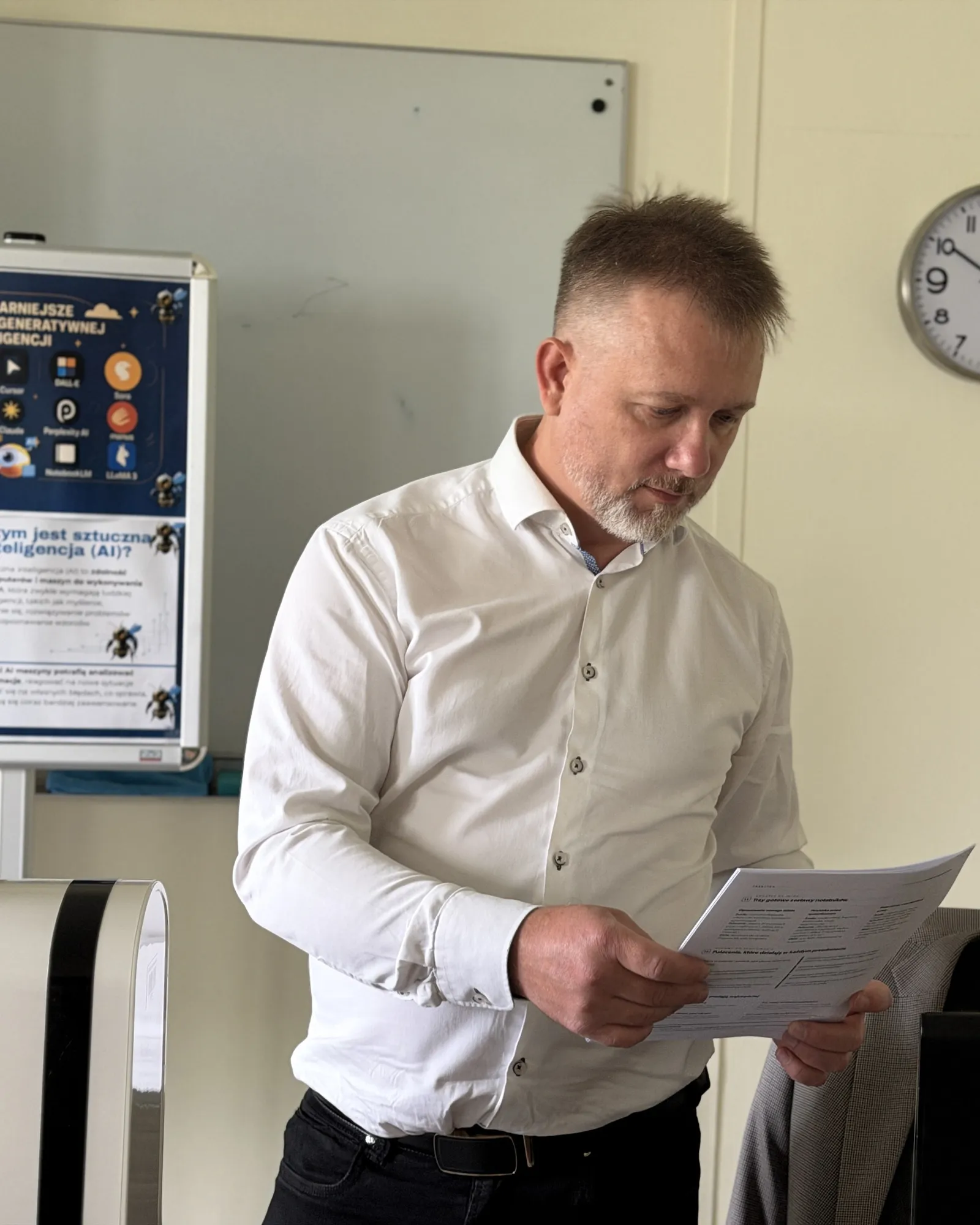 Piotr Kruk w białej koszuli czyta wydruk w szkolnej pracowni komputerowej, za nim plakat o narzędziach sztucznej inteligencji i zegar