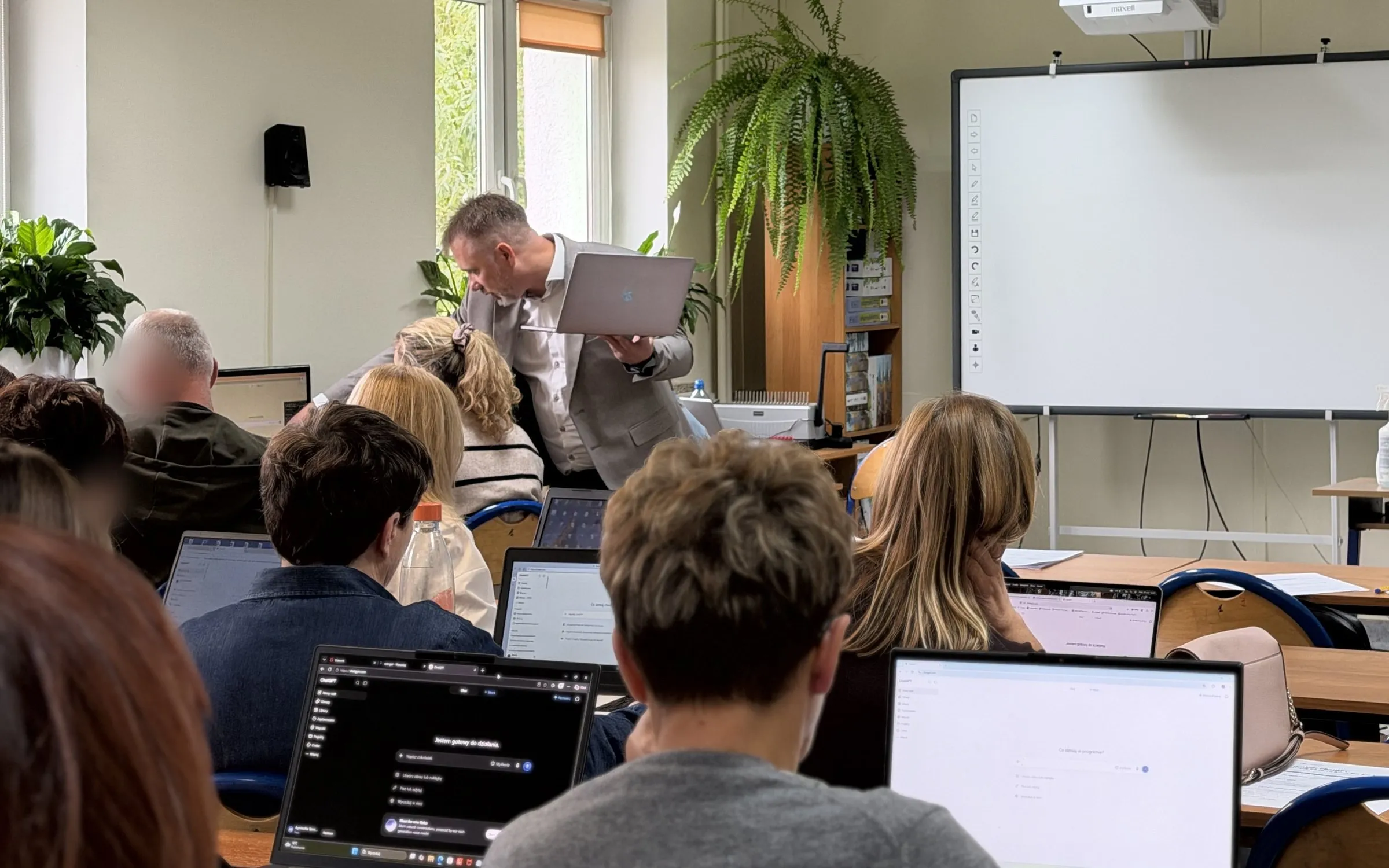 Piotr Kruk z laptopem w ręku pochyla się nad uczestniczką szkolenia i pomaga jej przy komputerze; pozostali uczestnicy pracują przy laptopach