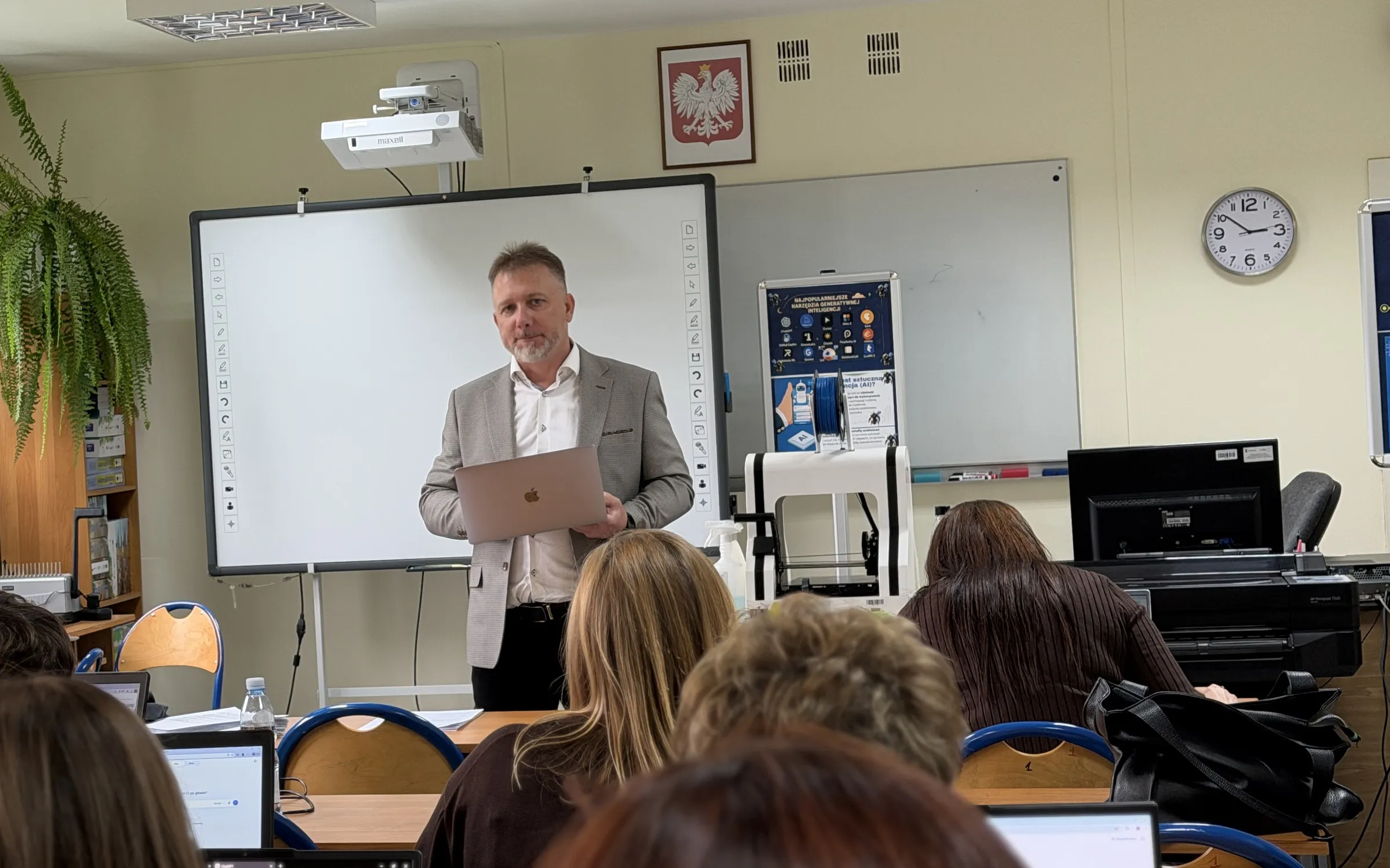 Piotr Kruk z laptopem prowadzi szkolenie z AI dla nauczycieli w szkolnej pracowni komputerowej; uczestnicy siedzą przy laptopach tyłem do obiektywu