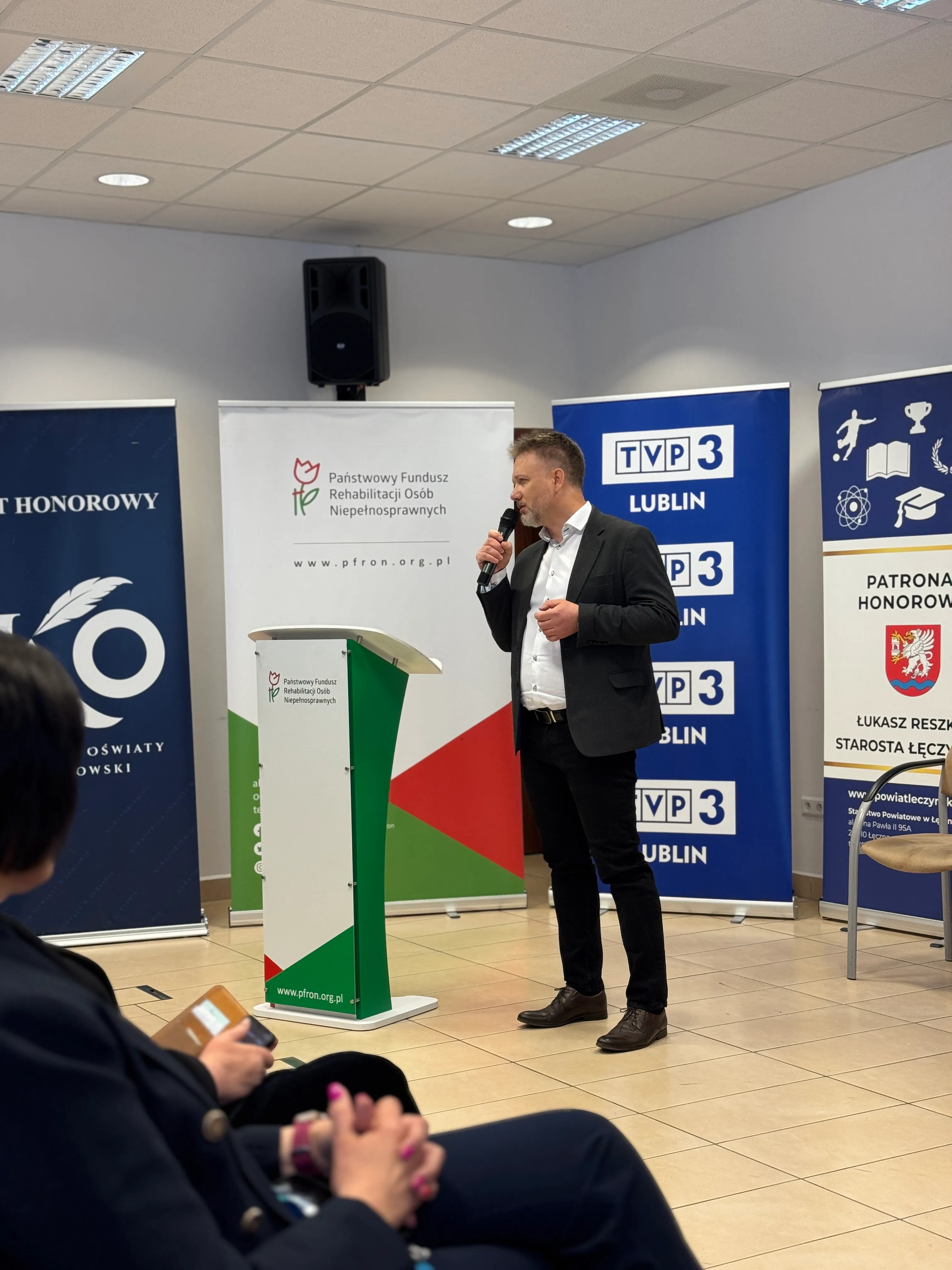 Piotr Kruk z mikrofonem podczas prelekcji o AI; w tle ścianki Kuratorium Oświaty, PFRON, TVP3 Lublin i patronatu honorowego Starosty Łęczyńskiego