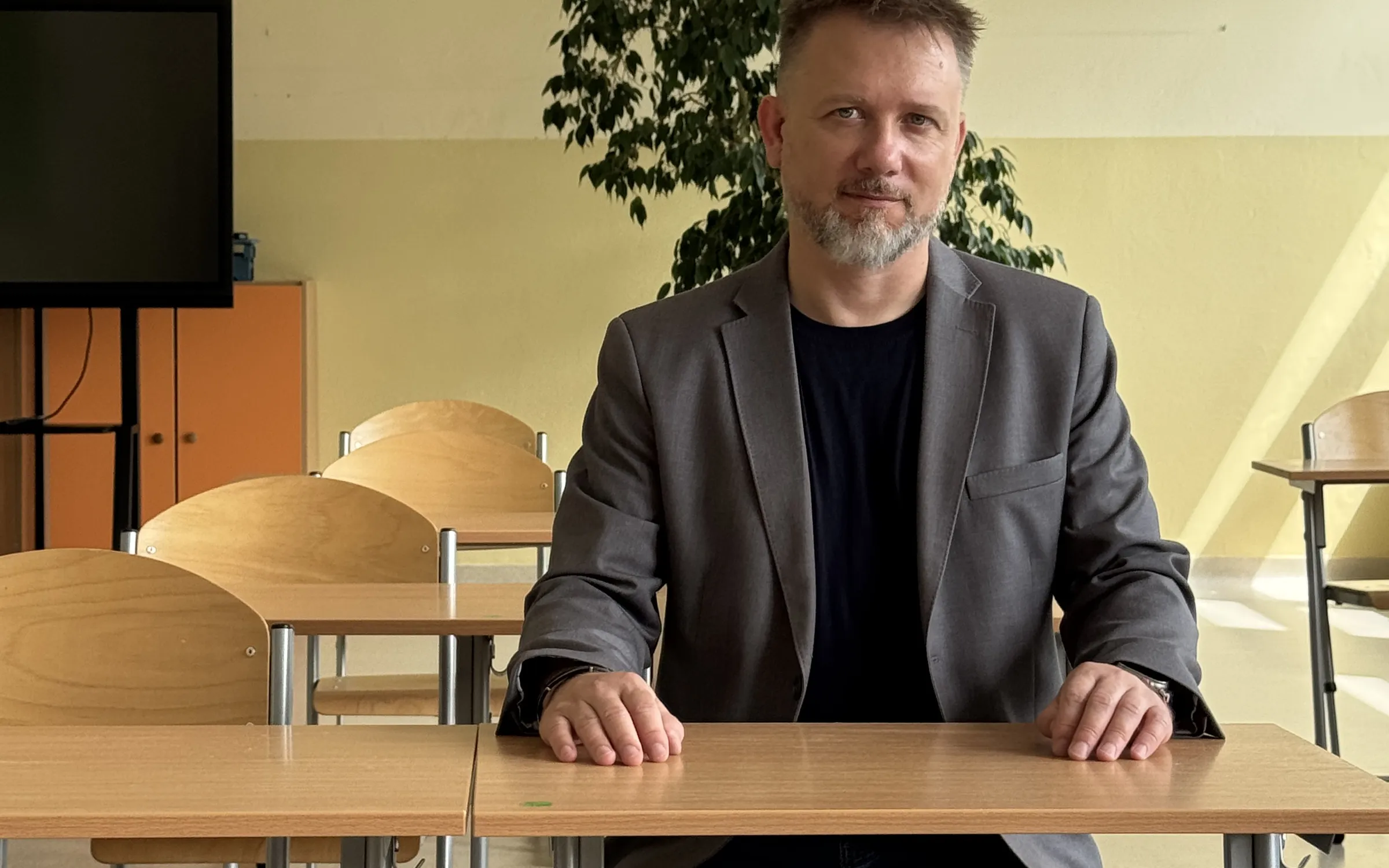 Piotr Kruk siedzi przy szkolnej ławce, z dłońmi opartymi o blat, i patrzy w obiektyw