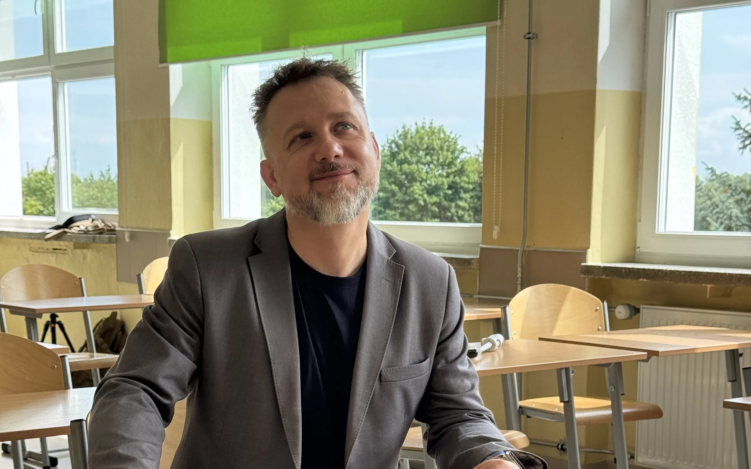 Piotr Kruk siedzi przy szkolnej ławce w pustej sali lekcyjnej, przy oknach z zielonymi roletami