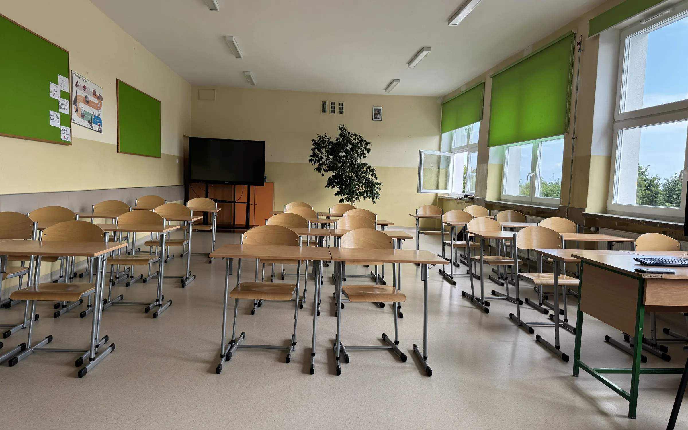 Pusta sala lekcyjna z rzędami ławek, tablicą i oknami z zielonymi roletami