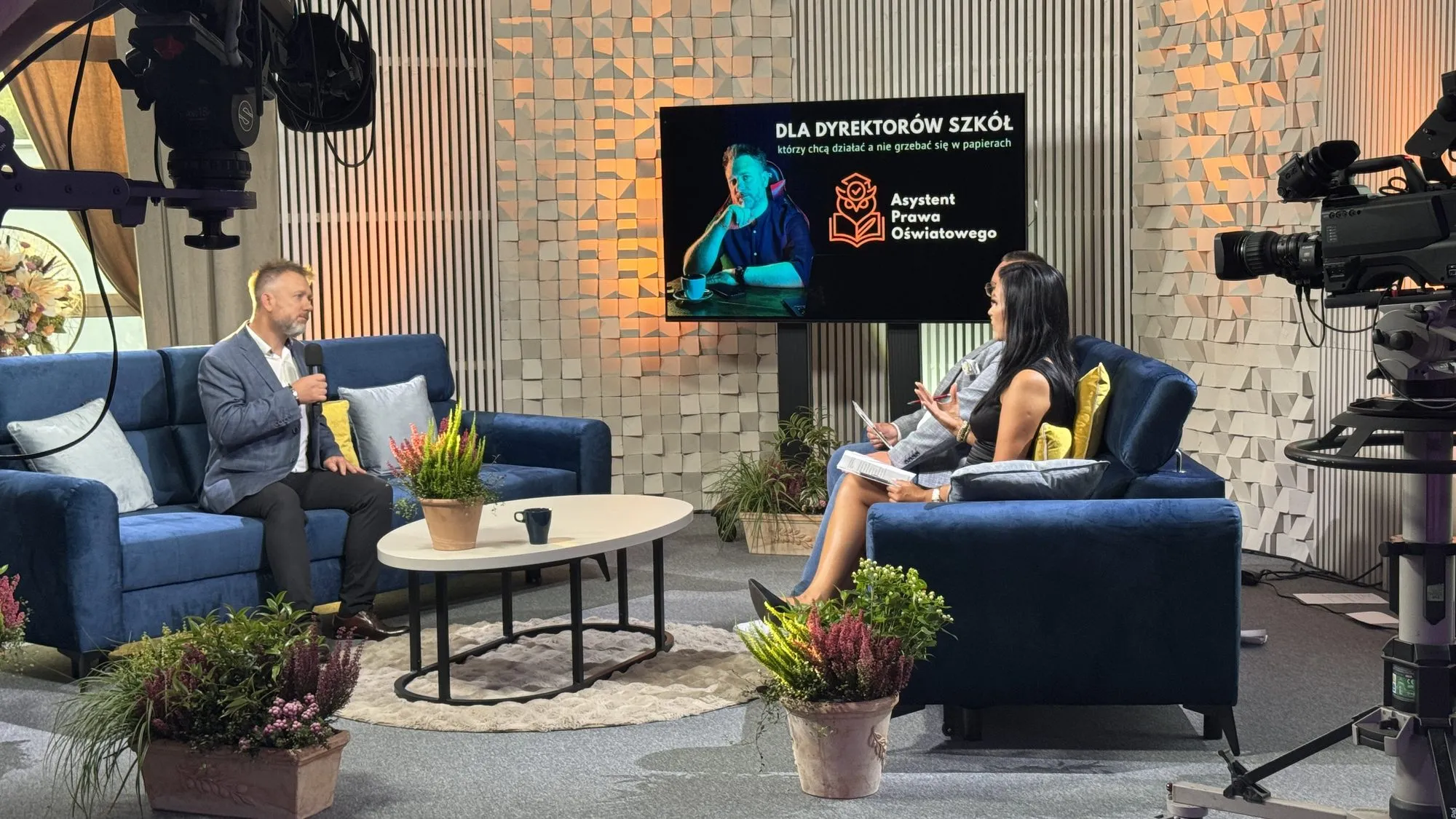 Studio telewizyjne podczas nagrania: Piotr Kruk z mikrofonem rozmawia z prowadzącą, na ekranie w tle plansza „Dla dyrektorów szkół — Asystent Prawa Oświatowego”, po bokach kamery