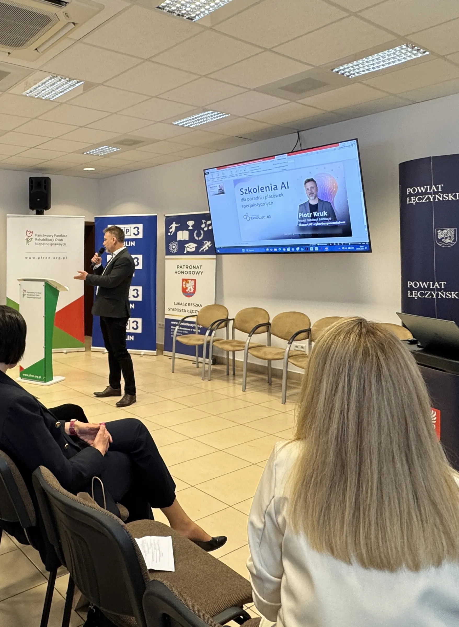 Piotr Kruk z mikrofonem prowadzi prelekcję dla poradni i placówek specjalistycznych; na ekranie tytuł szkolenia, w tle ścianki PFRON, Kuratorium Oświaty i Powiatu Łęczyńskiego