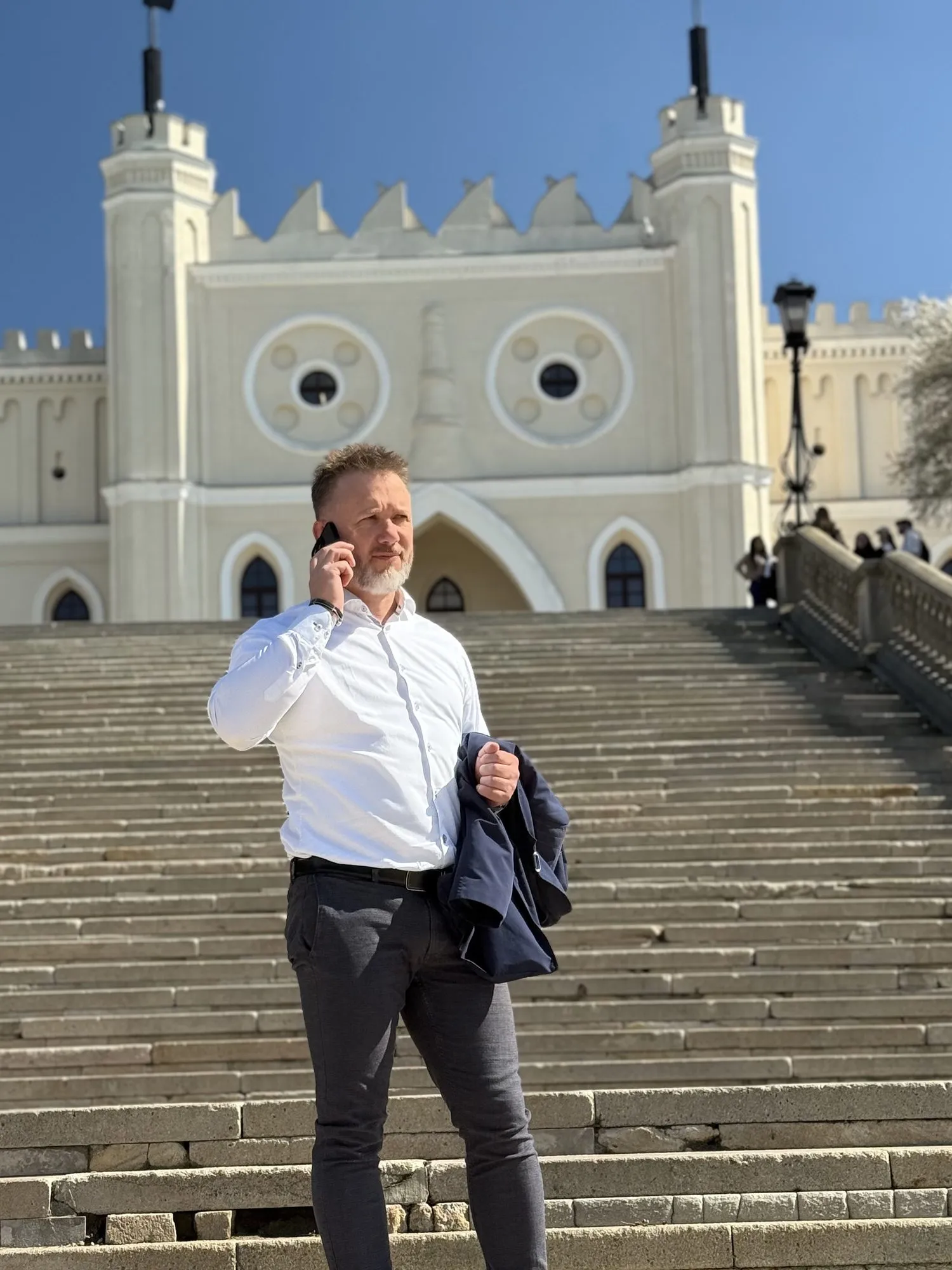 Piotr Kruk rozmawia przez telefon na schodach Zamku Lubelskiego w Lublinie, z marynarką przewieszoną przez ramię